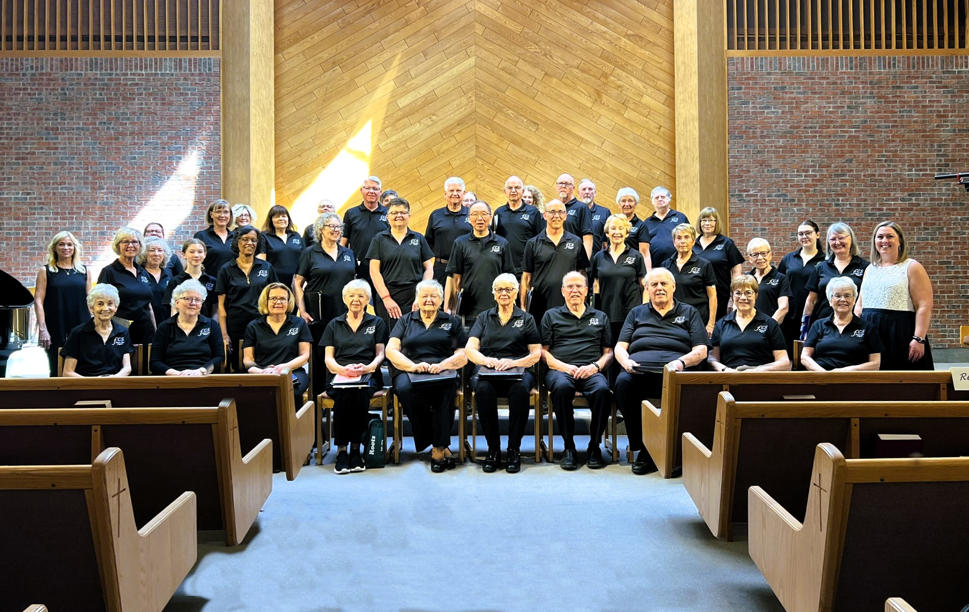 A group photo of the Forest City Singers, a community choir, standing and sitting in a church setting, wearing black choir shirts.